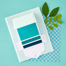 A layered paper craft or card design with white cardstock shapes and teal, aqua, and dark blue color blocks, placed on a turquoise background with a blue-and-white gingham sheet and green leaf cutouts.