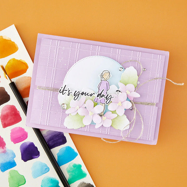 A handmade greeting card or craft paper piece on a solid orange background, styled with a watercolor paint palette and paintbrushes. The card is lavender purple with a grid texture, featuring a central white oval with a cartoon girl, the phrase 'it's your day' in black script, and layered pink and green paper flowers.