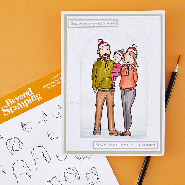 A greeting card featuring a hand-drawn family of four wearing red Santa hats, displayed on an orange textured background alongside a stamp booklet and a pencil.