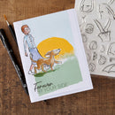 Card with illustration of a person and dog walking, surrounded by art supplies on a wooden surface.