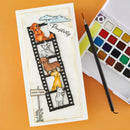 A handmade dog-themed card featuring a film strip design with illustrated dogs, a small sign reading 'Good Times,' and a 'Paw-activity' tag, displayed next to a watercolor paint palette and paintbrush on an orange background.