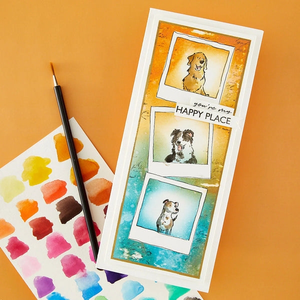 A handmade rectangular card featuring three watercolor dog portraits in polaroid-style frames with the text 'you're not HAPPY PLACE' across the center, styled on an orange background next to a paintbrush and a watercolor palette sheet.