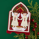 A decorative Christmas greeting card featuring two angel figures in white dresses holding red candles, set against a red and cream arched background with green leaves and gold stars. The card is displayed on a dark green background with artificial evergreen branches and red berries.