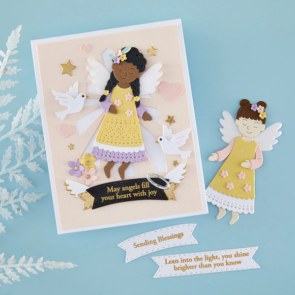 A handmade paper angel greeting card and matching cutout on a light blue background. The card features a layered angel figure in a yellow dress with white wings, surrounded by white doves, pink hearts, gold stars, and a black banner with blessings.