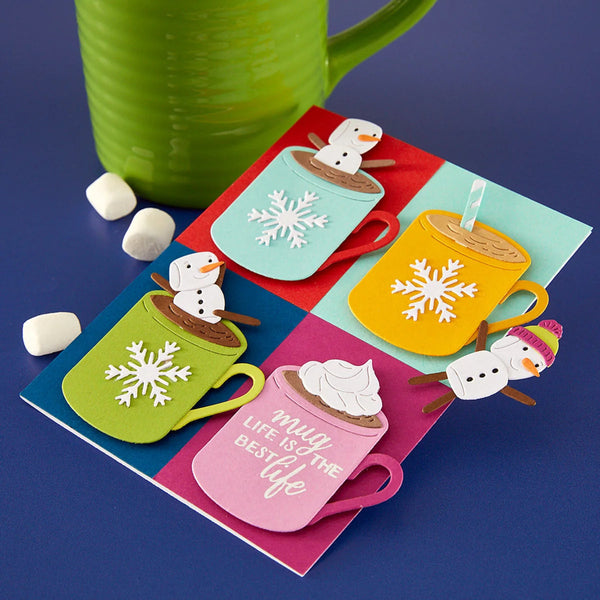 A colorful layered paper craft card featuring four hot cocoa cups with snowflakes and snowmen, displayed on a blue surface with a green mug and marshmallows.