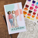 A handmade greeting card featuring an illustration of two children standing together, with the text 'off to great places' at the bottom. The card is displayed on a wooden surface alongside a paintbrush, a sheet of watercolor paint swatches, and a clear stamp sheet with face outlines.