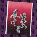 A Halloween greeting card featuring two dancing skeletons with the text 'YOU'RE BAD TO THE BONE' on a red-to-black gradient background, displayed against a purple patterned backdrop of skulls and hearts.