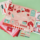 A flat-lay display of a 'Dear Santa' spelling out kit by Spellbinders, featuring a pink illustrated box surrounded by red, green, and white craft sheets, stickers, and small cards on a mint green background.
