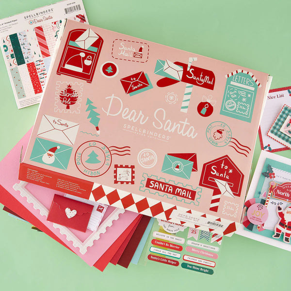 A flat-lay display of a 'Dear Santa' spelling out kit by Spellbinders, featuring a pink illustrated box surrounded by red, green, and white craft sheets, stickers, and small cards on a mint green background.