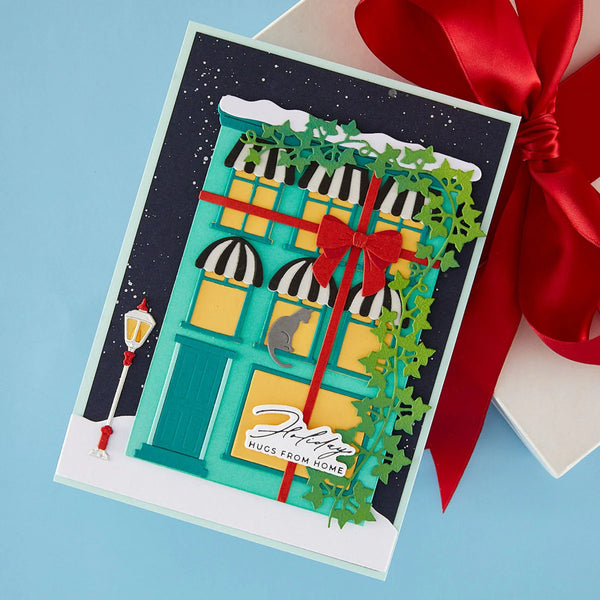 A handmade layered paper greeting card featuring a festive holiday storefront design, styled on top of a gift box with a red ribbon against a light blue background.