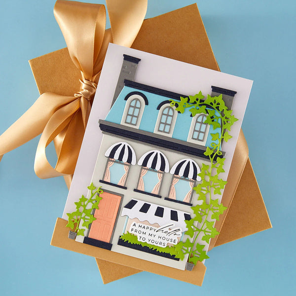 A handmade greeting card featuring a detailed illustration of a house with black-and-white awnings, green ivy, and the text 'A Happy little House from my house to yours,' styled on top of a kraft envelope with a gold ribbon against a light blue background.
