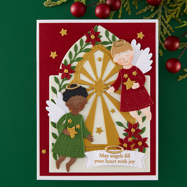 A layered Christmas greeting card featuring two angel figures, one with dark skin and one with light skin, both wearing red and green dresses and surrounded by red flowers, green leaves, and a gold starburst. The card is displayed on a dark green background with red ornaments and pine branches.