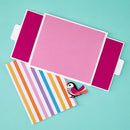 A pink rectangular card or paper insert with white borders and magenta side tabs, laid on a turquoise background. A striped paper sheet with orange, yellow, blue, purple, and white stripes lies beneath it, with a small paper bird decoration resting on top.
