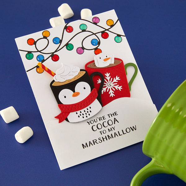 A handmade layered paper card featuring penguin cocoa cups under colorful string lights, with the text 'YOU'RE THE COCOA TO MY MARSHMALLOW', displayed on a blue background with marshmallows and a green mug prop.