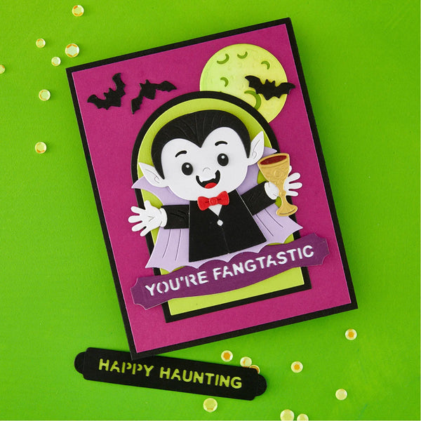 A handmade Halloween greeting card featuring a cartoon vampire holding a gold goblet, with black bats, a yellow moon, and the text 'YOU'RE FANGTASTIC' on a bright green background. A small black tag below reads 'HAPPY HAUNTING'.