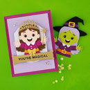 A handmade layered paper craft set featuring a purple card with a cartoon princess and the words 'YOU'RE MAGICAL' next to a matching witch character cutout, displayed on a bright green background.