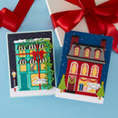 Two handmade Christmas greeting cards displayed on a light blue background with a white gift box and red ribbon partially visible behind them. The left card shows a turquoise storefront decorated with green ivy and a red bow, while the right card shows a red house with yellow windows under a dark blue starry sky.