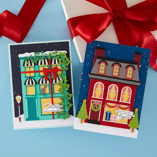 Two handmade Christmas greeting cards displayed on a light blue background with a white gift box and red ribbon partially visible behind them. The left card shows a turquoise storefront decorated with green ivy and a red bow, while the right card shows a red house with yellow windows under a dark blue starry sky.