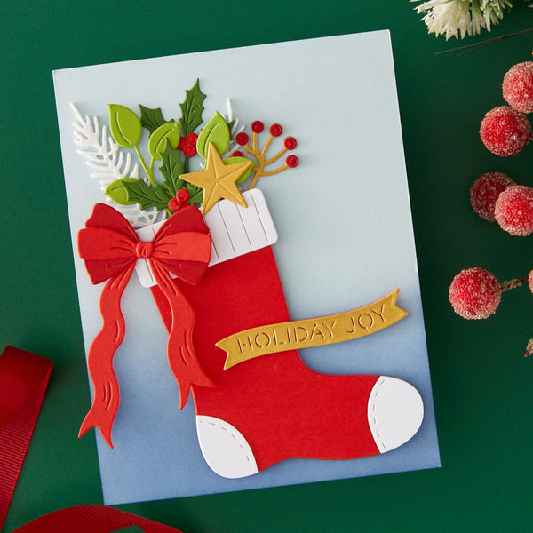 A handmade layered Christmas stocking card on a light blue cardstock base, decorated with red felt stocking, green holly leaves, red berries, a gold star, a red bow, and a gold 'HOLIDAY JOY' banner, photographed on a dark green background with decorative red berries and white flowers.