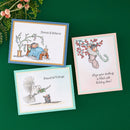 Three illustrated greeting cards arranged on a dark green background with evergreen branches and berries at the top left. The cards feature whimsical holiday illustrations: a sleeping figure under a tree reading 'Dream & Believe,' a white bird carrying a green sprig with 'Peaceful Tidings,' and a teddy bear under berry branches with a message about stocking.