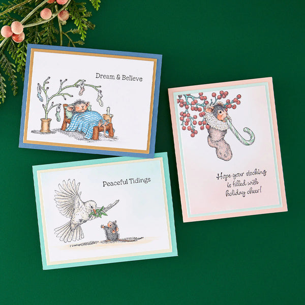 Three illustrated greeting cards arranged on a dark green background with evergreen branches and berries at the top left. The cards feature whimsical holiday illustrations: a sleeping figure under a tree reading 'Dream & Believe,' a white bird carrying a green sprig with 'Peaceful Tidings,' and a teddy bear under berry branches with a message about stocking.