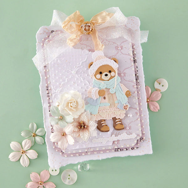 A decorative handmade card featuring a teddy bear illustration in winter clothing, embellished with layered paper, fabric flowers, pearls, and a ribbon bow, set against a pale mint green background with scattered floral embellishments.
