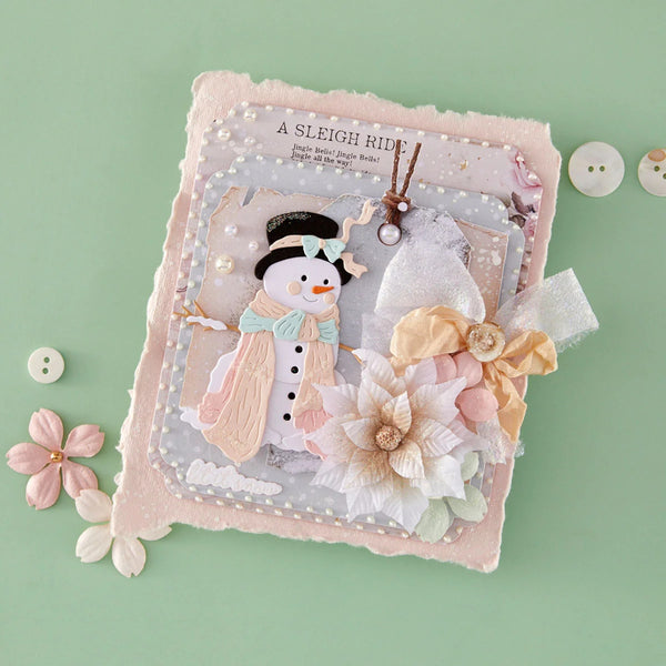 A handmade winter-themed scrapbook or journal page featuring a 3D snowman, layered paper flowers, and a pink textured border, styled on a mint green background with decorative buttons.