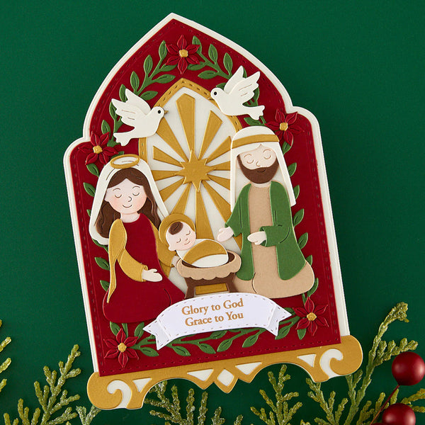 A decorative paper-cut style Christmas nativity sign featuring Mary, Joseph, baby Jesus, and a dove, with the text 'Glory to God Grace to You' on a green background with holiday greenery and red ornaments.