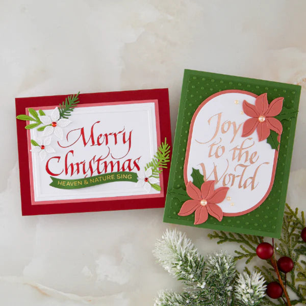 Two handmade Christmas greeting cards displayed on a light marble surface with holiday greenery and red berries. One card reads “Merry Christmas” in red script on a white background with a red border, and the other reads “Joy to the World” in gold script on a green polka-dot background with layered coral flowers and green leaves.