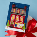 A Christmas-themed greeting card featuring a red house with yellow windows, decorated with a wreath and small trees, shown resting on a white gift box tied with a red ribbon against a light blue background.