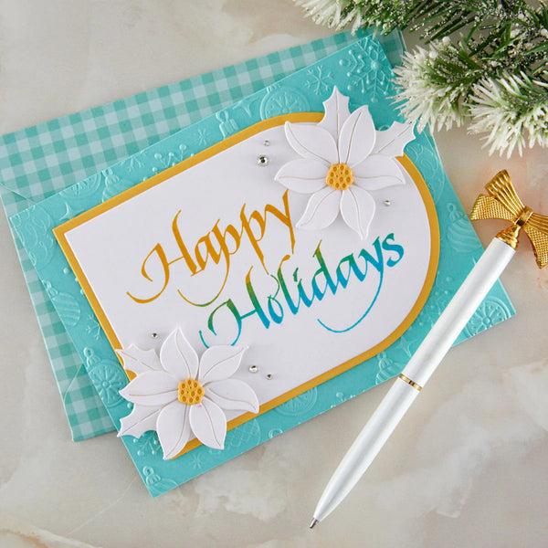 A handmade holiday greeting card with 'Happy Holidays' in gold and teal script, decorated with two white daisy flowers, shown on a turquoise gingham background next to a white and gold pen.