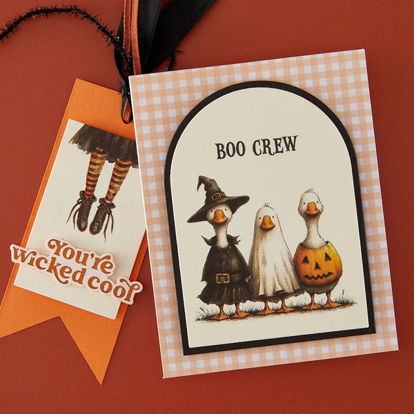 A Halloween-themed greeting card and matching tag on a warm orange-brown background. The main card has a gingham border and an arched illustration of three cartoon geese dressed as witch, ghost, and pumpkin.