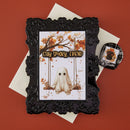 An ornate black picture frame holding a Halloween greeting card with a ghost on a swing and the words 'STAY SPOOKY, FRIEND,' styled on top of white envelopes against a terracotta background. A small matching black-and-white accessory piece with a spiderweb design and black cat illustration sits to the right.