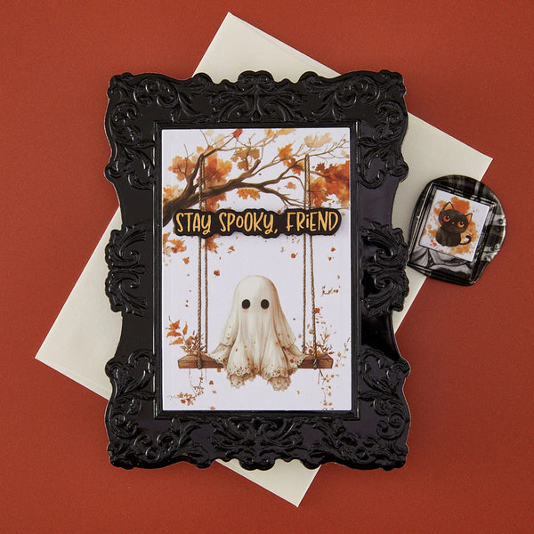 An ornate black picture frame holding a Halloween greeting card with a ghost on a swing and the words 'STAY SPOOKY, FRIEND,' styled on top of white envelopes against a terracotta background. A small matching black-and-white accessory piece with a spiderweb design and black cat illustration sits to the right.