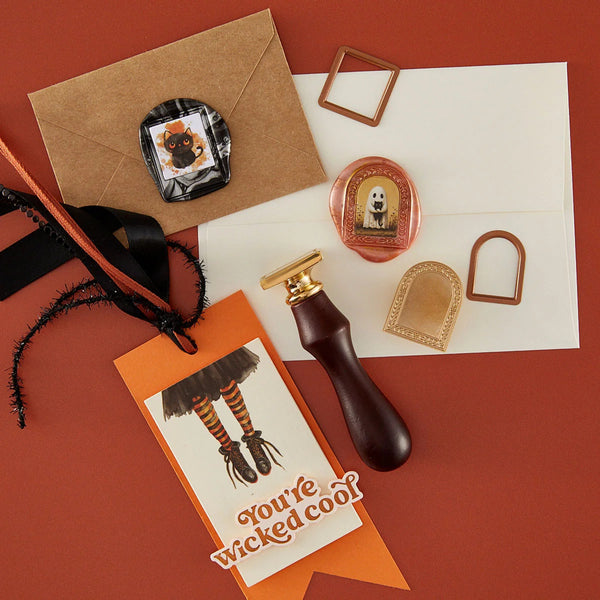 A flat-lay of a Halloween-themed stationery set on a terracotta background, featuring a brown kraft envelope, a white card with a ghost illustration, an orange tag reading 'You're Wicked cool!', a dark brown leather seal stamp handle, gold metal frame pieces, and a round pink wax seal.