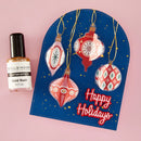 A flat-lay of a Spellbinders 'Gold Rush' glitter nail polish bottle next to a blue arched holiday card with red and white ornament illustrations and 'Happy Holidays' text, set against a solid pink background.