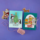 A flat lay of handmade Christmas greeting cards and decorative stickers on a purple background. The set includes a light blue card with penguins by a Christmas tree, a teal card with festive animals and the words 'Season'sreetings', a Santa sticker, a dachshund sticker, and a spool of red-and-white twine.
