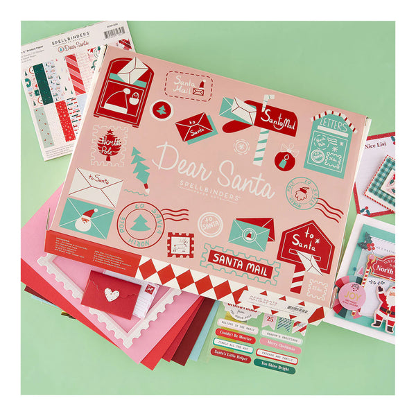 A flat-lay display of a Spellbinders Dear Santa Spelling Activity Kit, featuring a pink cover with red and teal holiday illustrations, surrounded by matching red, pink, and teal paper sheets and sticker sheets on a mint green background.