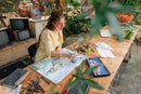 A woman sits at a wooden table outdoors, painting on a large canvas with colored pencils and art supplies around her. The scene is styled with vintage TVs, plants, and foliage in the background.