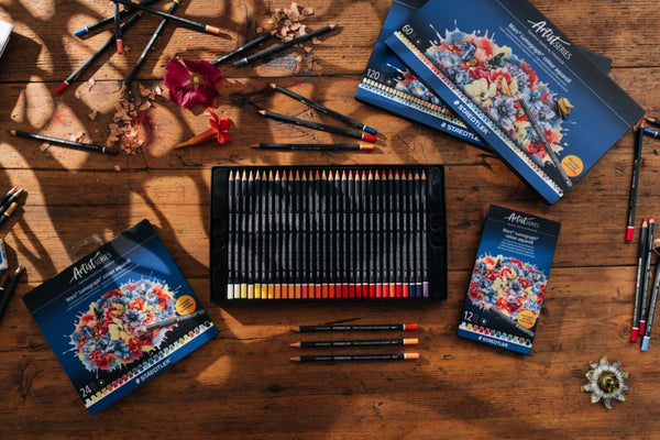 An ArtFinesse colored pencil set arranged on a rustic wooden table, with the open tray of black pencils in the center, matching product boxes around it, and several loose colored pencils placed around the scene. The boxes feature a colorful floral illustration and the ArtFinesse brand name.