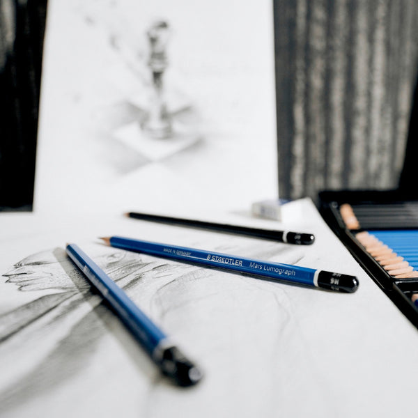 Staedtler drawing pencils on a white surface with blurred artwork in the background