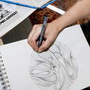 Person drawing in a sketchbook with a pen, surrounded by art supplies.