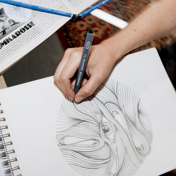 Person drawing in a sketchbook with a pen, surrounded by art supplies.