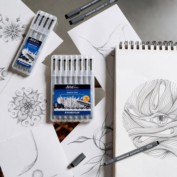 Art supplies including markers, pens, and a notebook with artistic drawings on a gray surface.