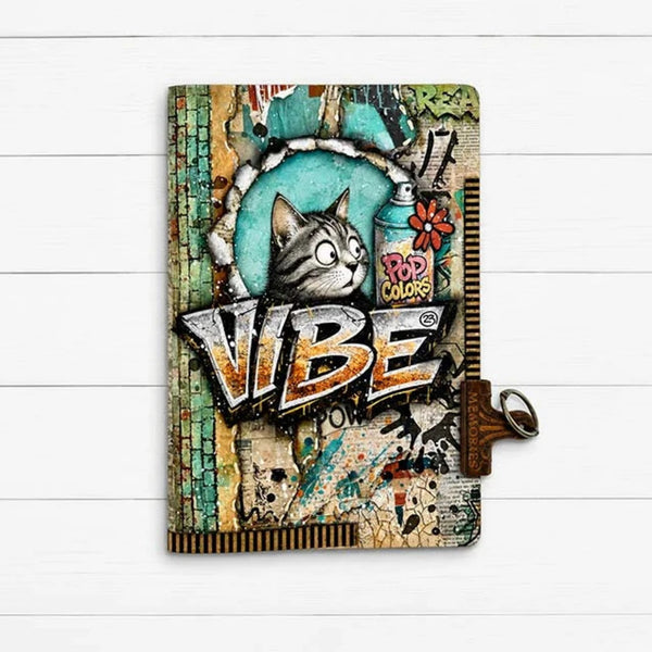 A hardcover notebook with a colorful graffiti-style cover featuring a cat illustration and the word 'VIBE' in large graffiti letters, shown on a white wood background with a brown strap and metal clasp.