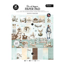 A die-cut designer paper pad titled 'Rustic Winter' by Studio Essentials, featuring a collage of winter-themed illustrations including skiers, snow dogs, deer, cabins, and patterned paper swatches in soft pastel tones.