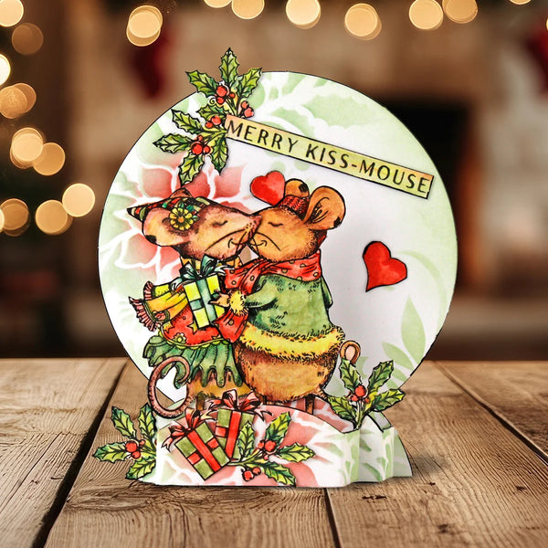 A round Christmas-themed decorative plaque or coaster featuring two kissing mice, wrapped gifts, red hearts, holly, and the text 'MERRY KISS-MOUSE' on a wooden tabletop with warm blurred lights in the background.