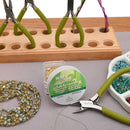 A bead craft setup on a gray surface with a spool of Bead Studio 21 Flex-Rite clear seed beads in the center, green-handled pliers, a wooden tool holder, and beads arranged in bowls in the background and foreground.
