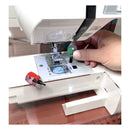 A close-up of a white sewing machine with a hand using a small green-handled screwdriver to remove a screw near the needle area. A second screwdriver with a red handle rests on the machine bed, and a wooden surface is visible underneath.