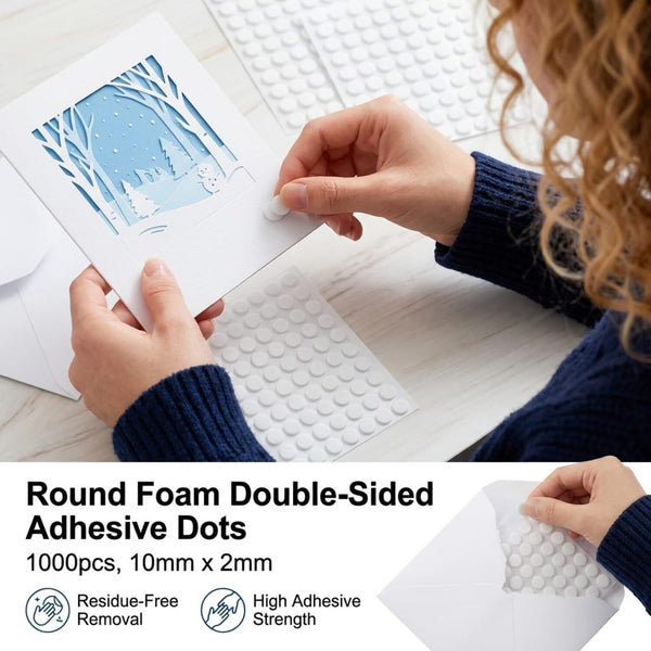 A person holds a white card decorated with layered winter trees and a snowy scene, while applying round adhesive dots from a sheet. The image includes text reading 'Round Foam Double-Sided Adhesive Dots, 1000pcs, 10mm x 2mm' with feature icons for residue-free removal and high adhesive strength.